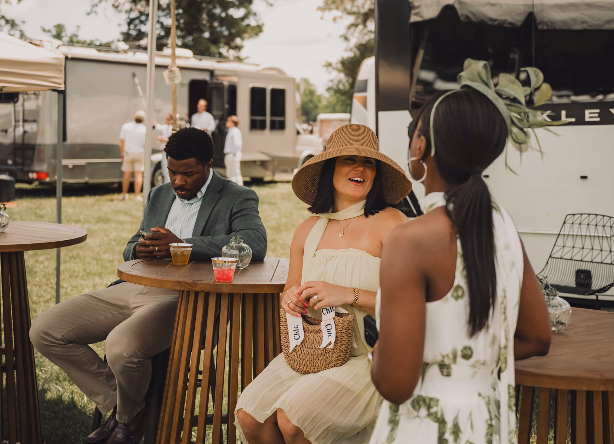 Two women chatting at outdoor event with man sitting nearby looking at phone, trailers in background.