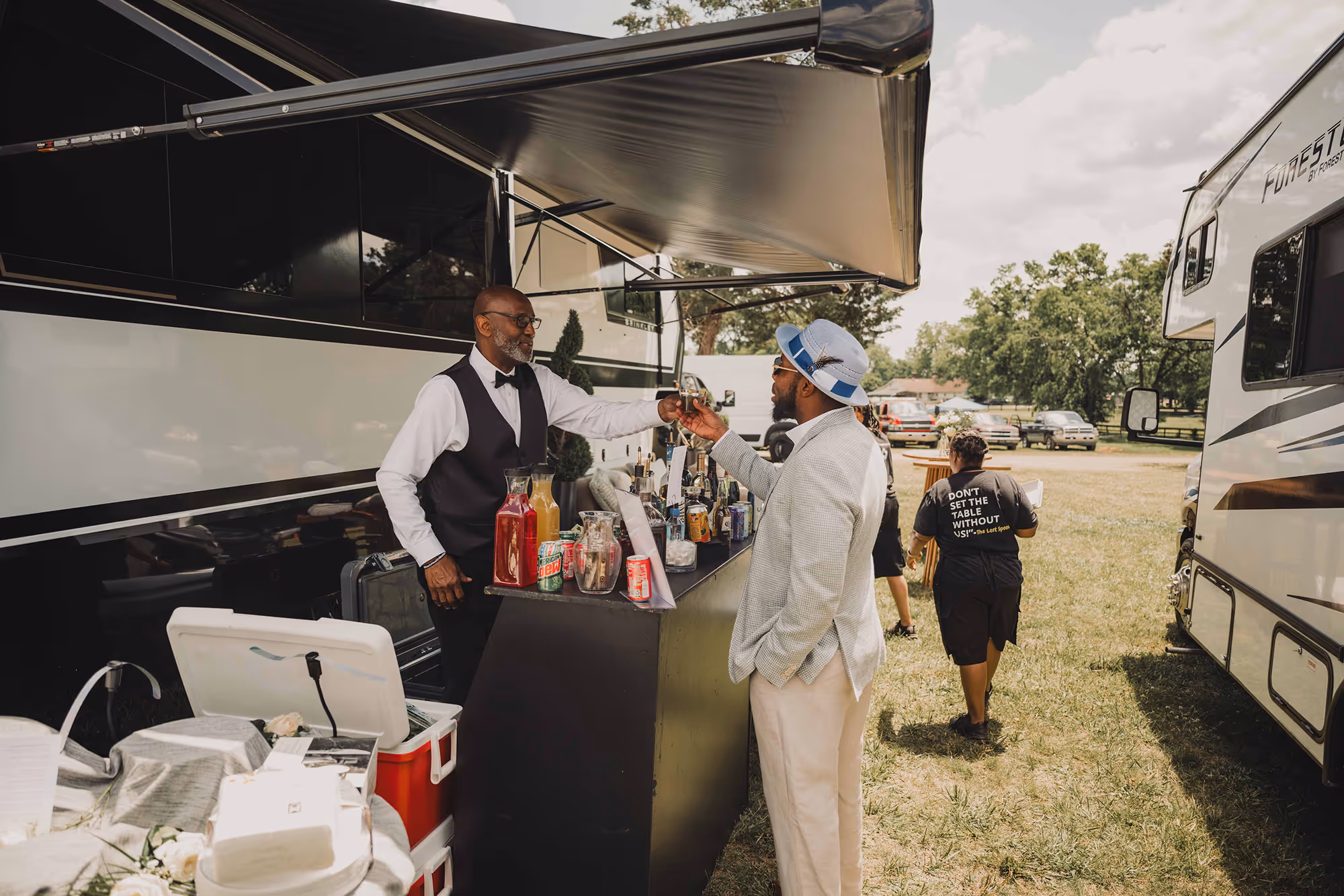 Bartender in vest and bow tie serving drink to man in hat and light jacket at outdoor bar near RVs.