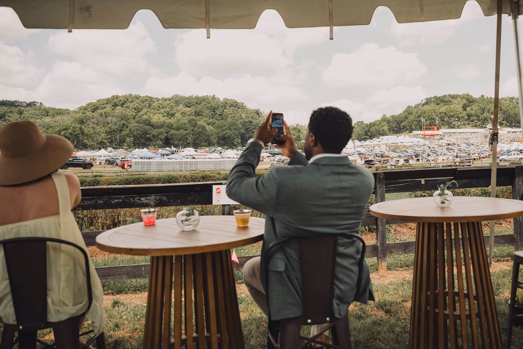People seated at wooden tables under a tent, one taking a photo of a large outdoor event in the distance.