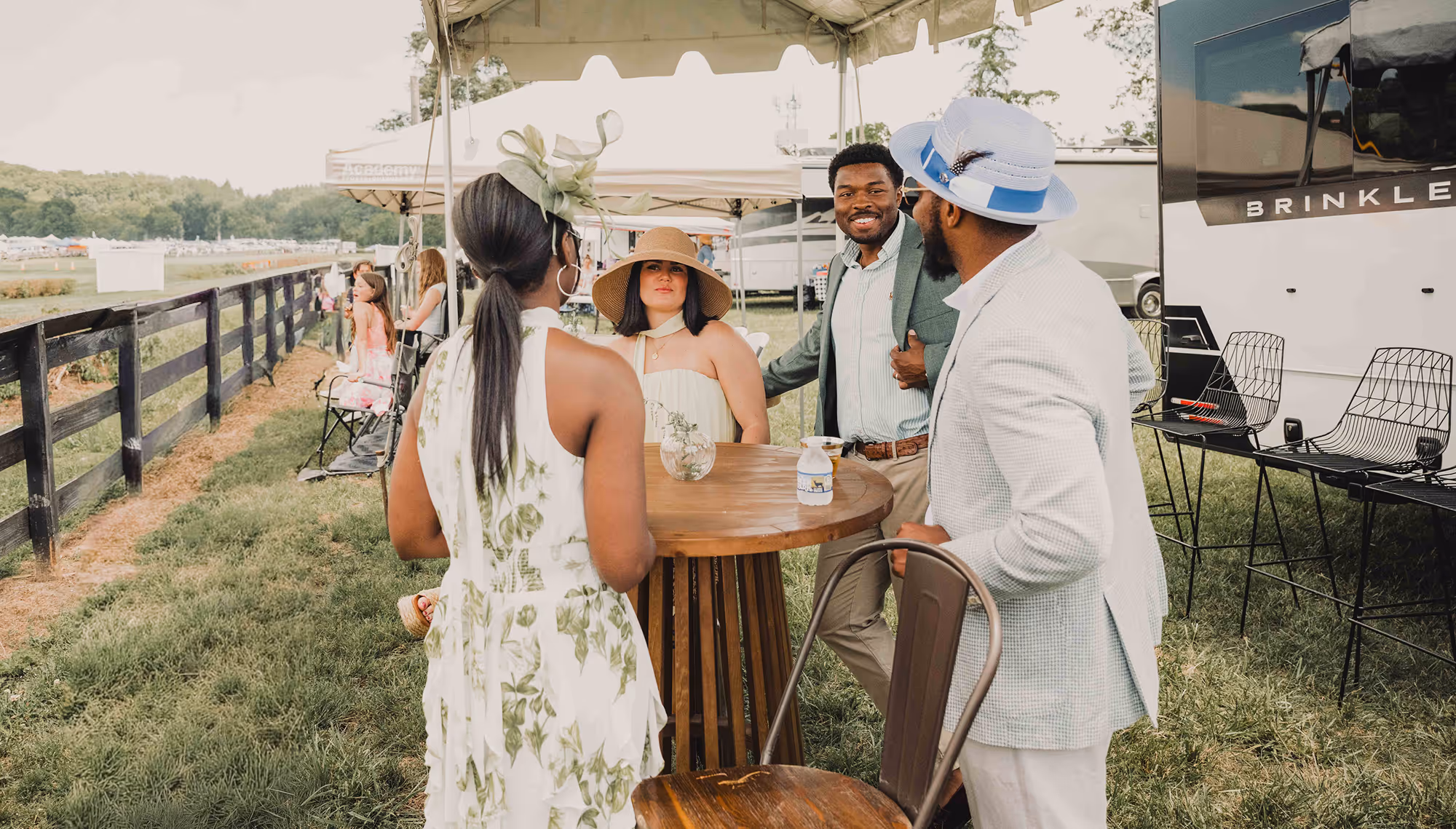 Four people in summer attire chatting outdoors around a high wooden table under a canopy.