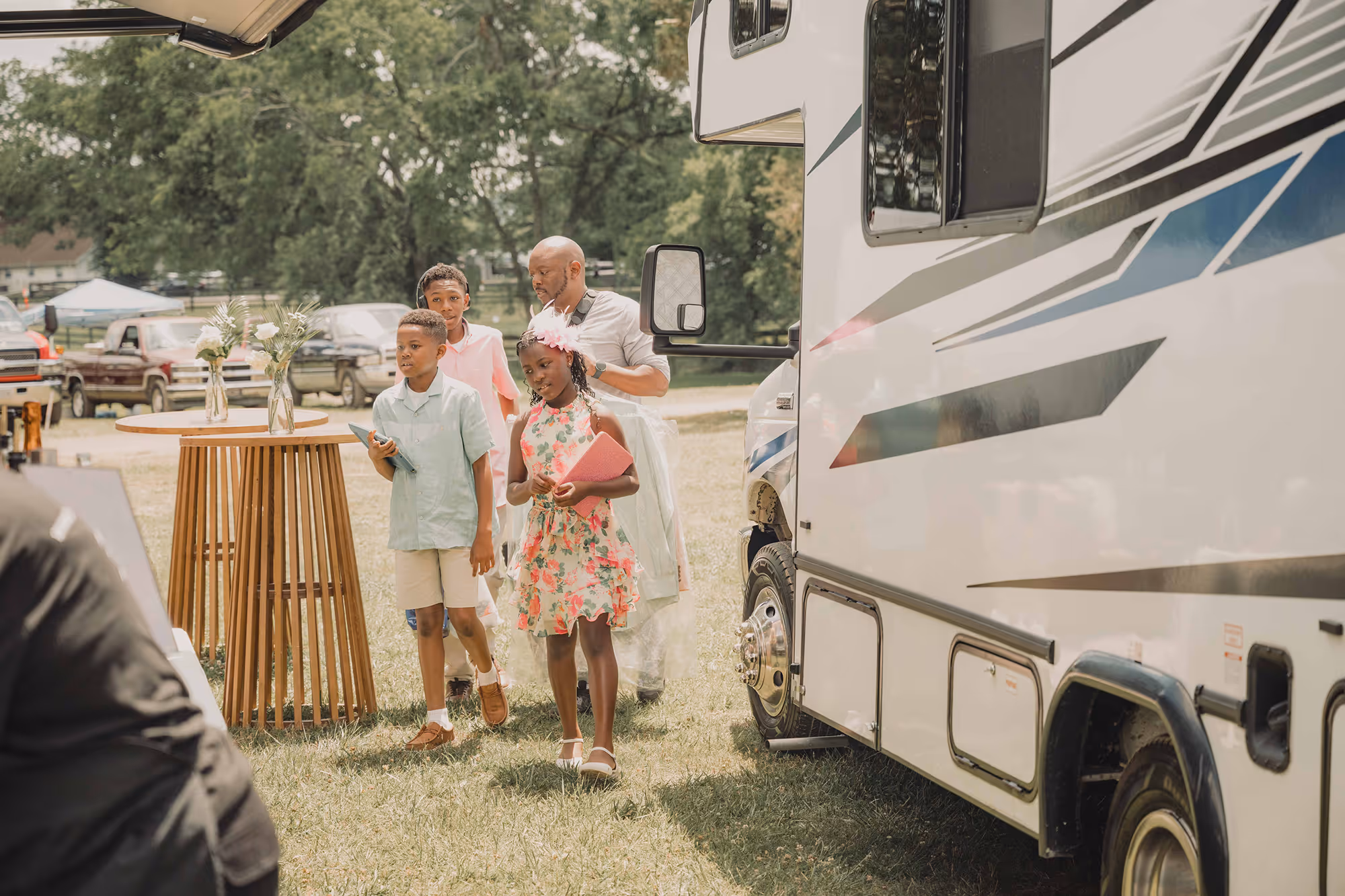 Man and three children holding folders walking on grass beside an RV during a daytime outdoor event.