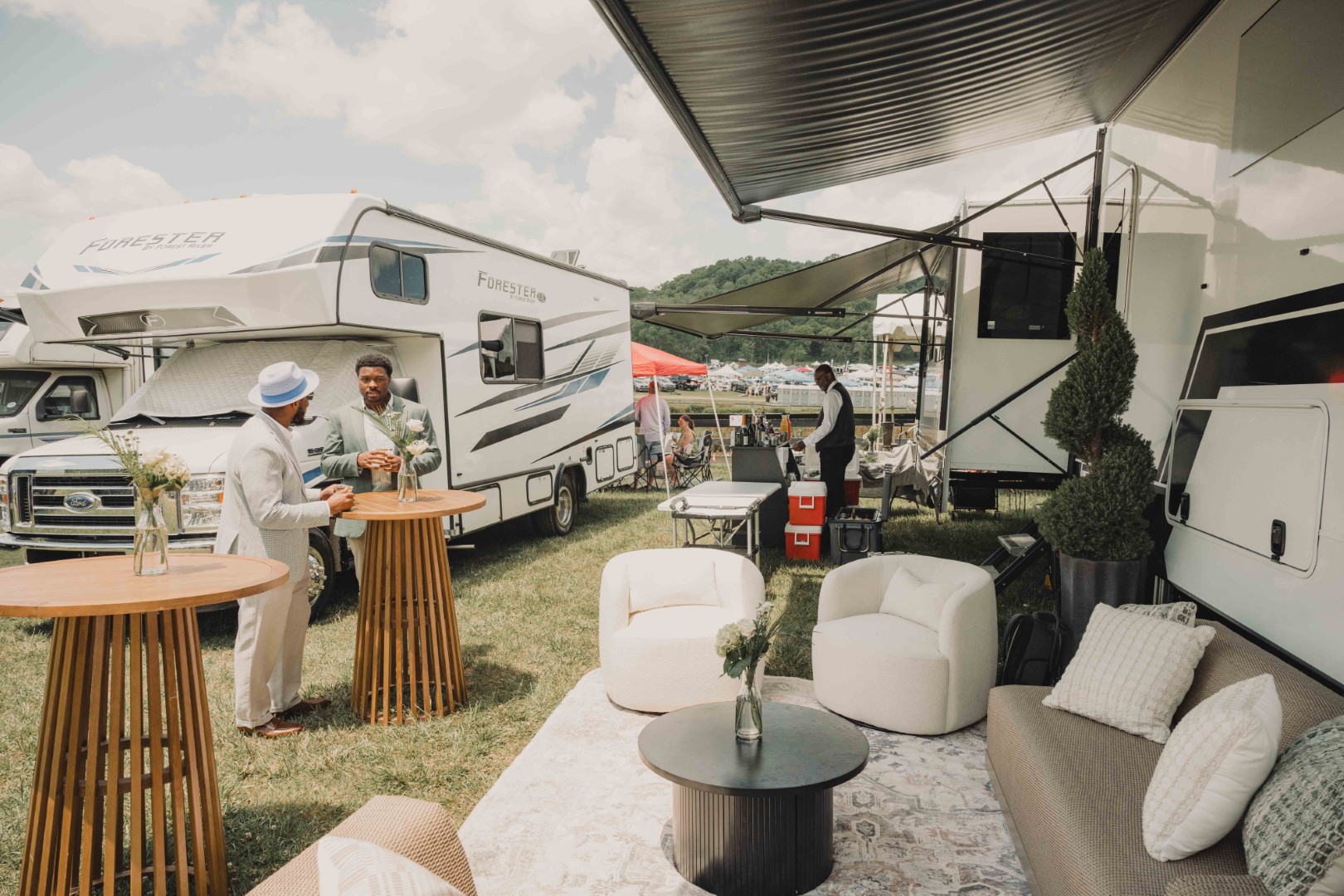 Two men talk at a wooden table near RVs with outdoor seating and a bartender in the background.