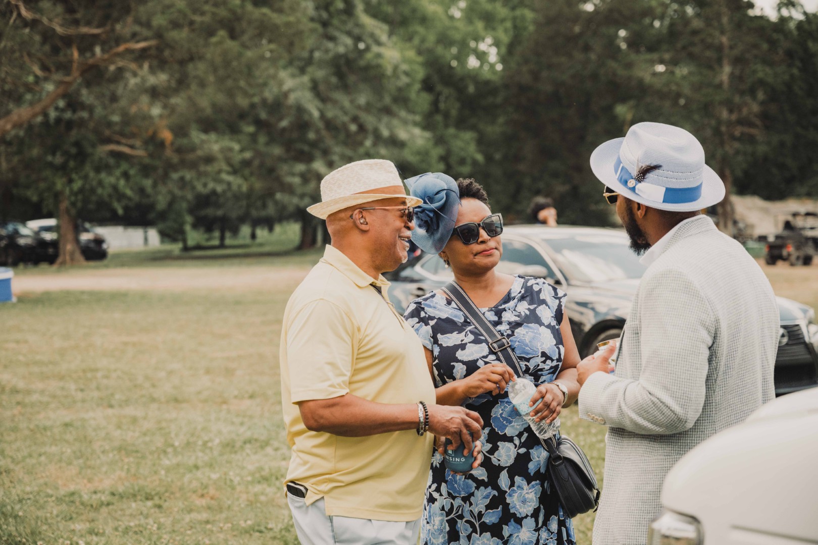 Three adults in hats talking outdoors near parked cars in a grassy, tree-filled area.