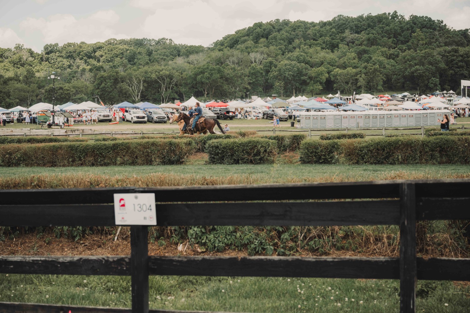Two riders on horseback near a fence with tents and portable toilets at a crowded outdoor event.