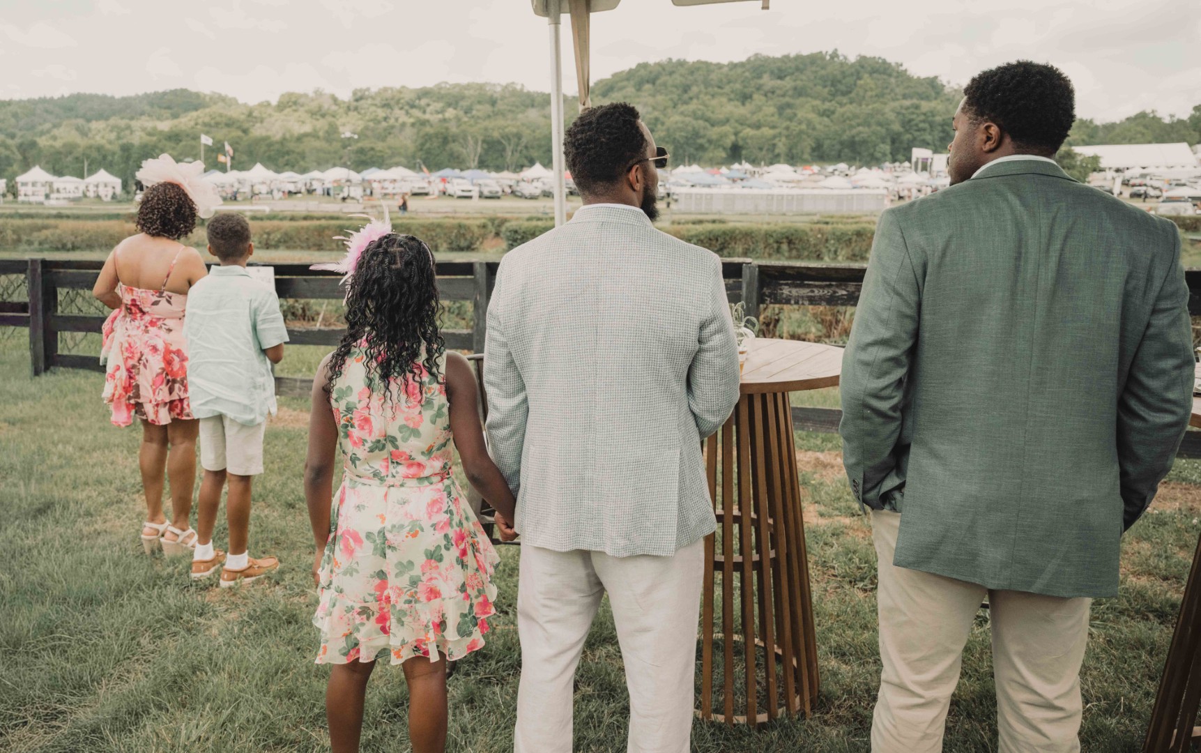 Five people in summer outfits stand with backs to camera near fence, watching an outdoor event.