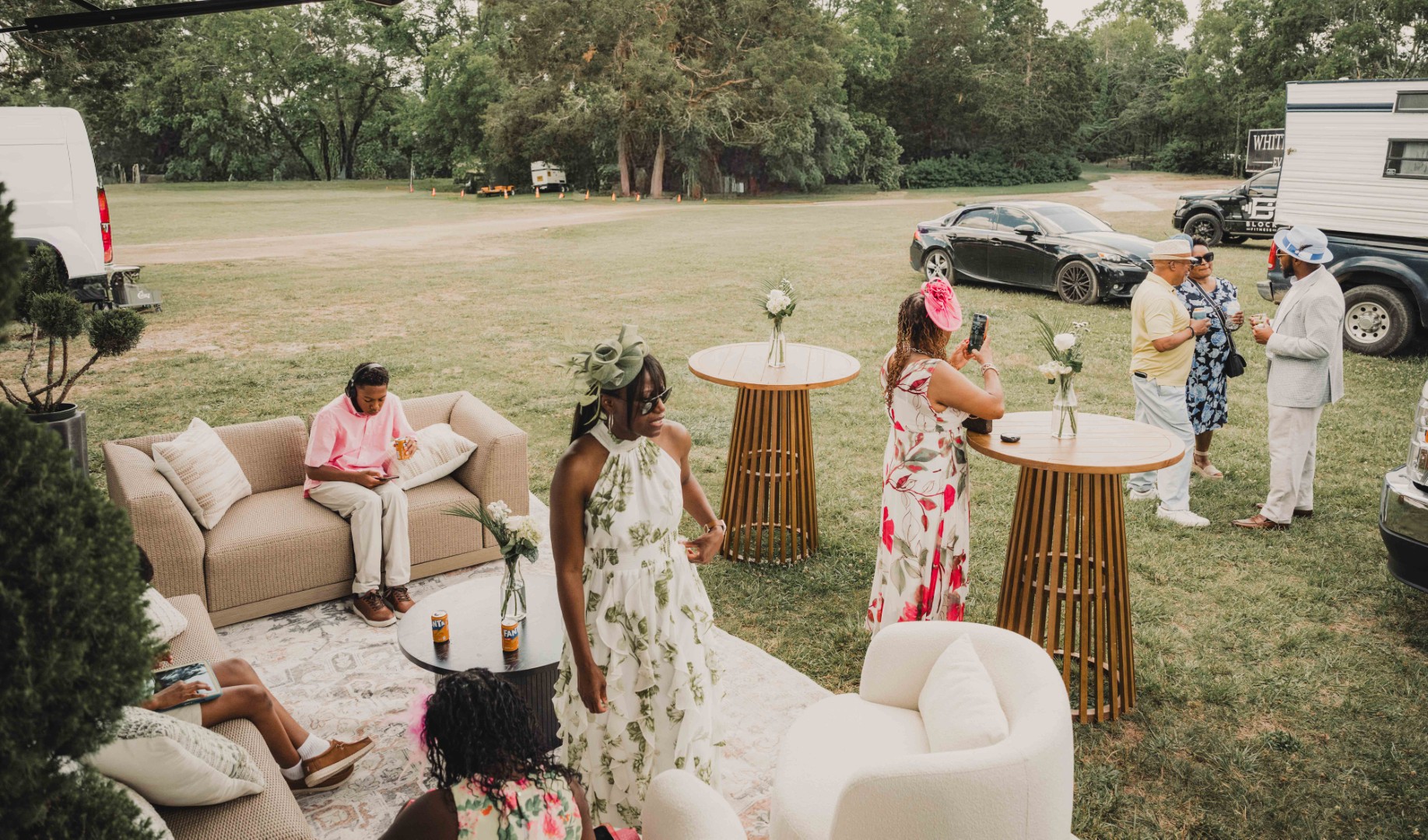 People socializing outdoors on grass with modern lounge furniture and wooden high tables with flowers.