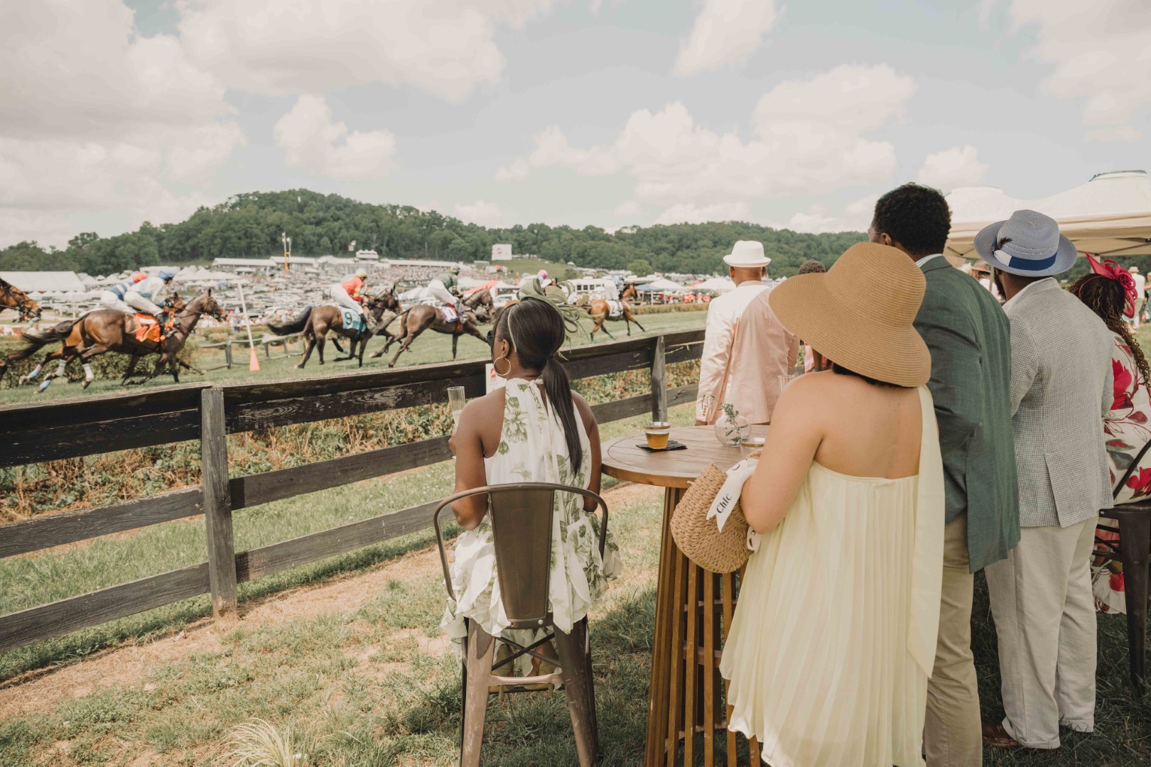 Group of well-dressed people watching a horse race at an outdoor event on a sunny day.