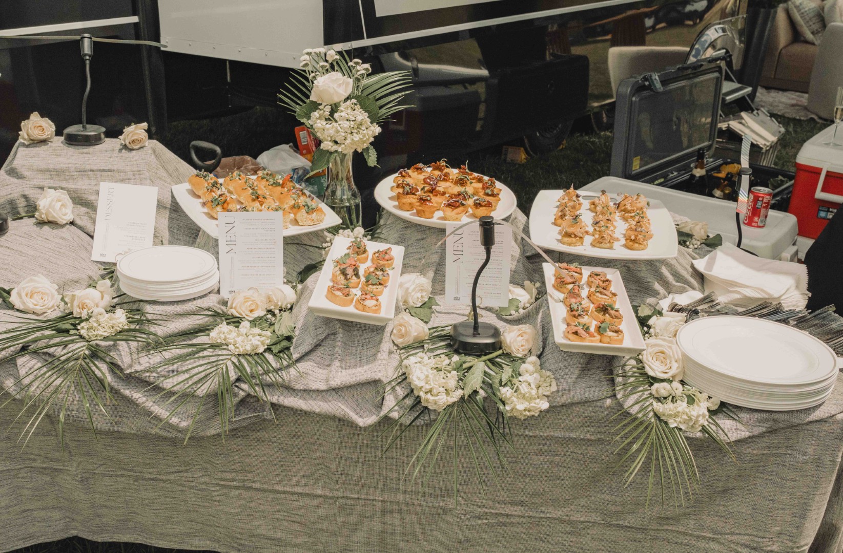 Elegant buffet table with assorted appetizers, floral decorations, and menus on linen cloth outdoors.