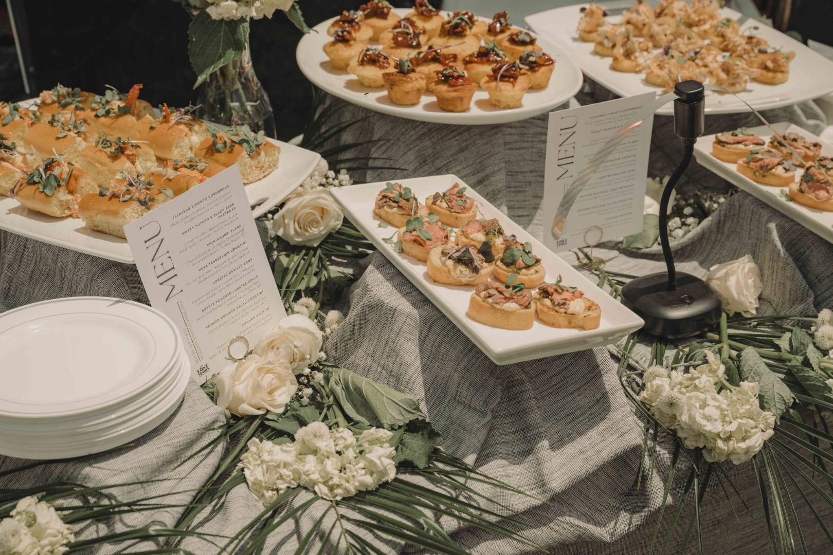 Long table with white tablecloth and empty dessert stands on green lawn under tall pine trees.