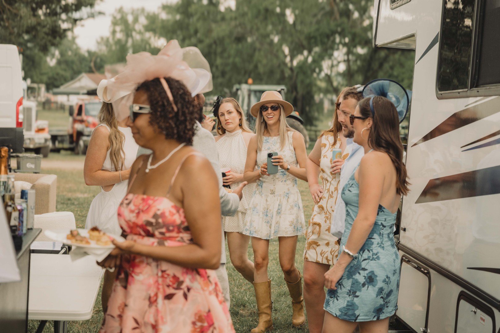 Group of people dressed in summer attire socializing outdoors near a camper and food table.