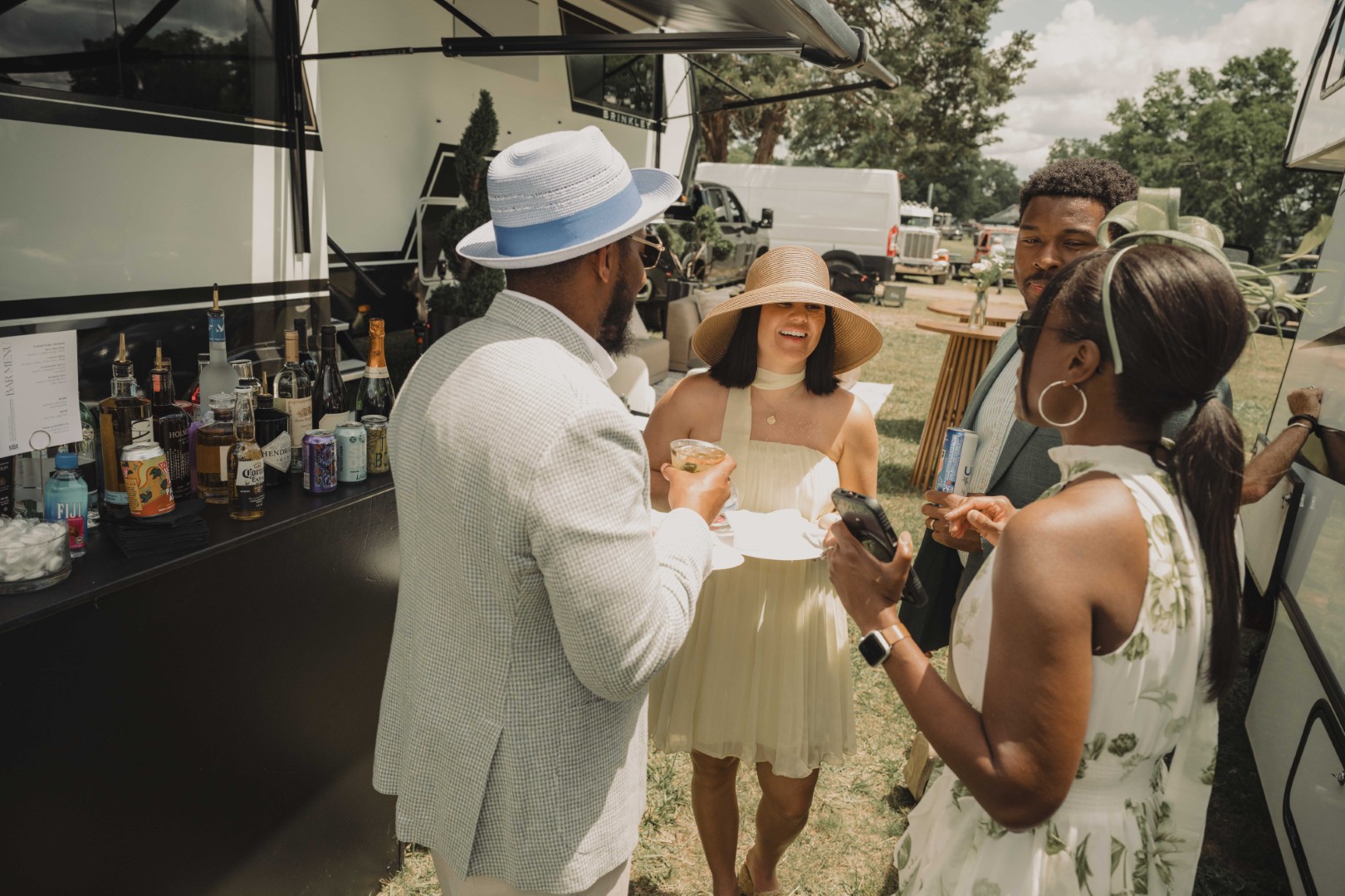 Four well-dressed people chat outdoors near a bar setup with drinks on a sunny day.