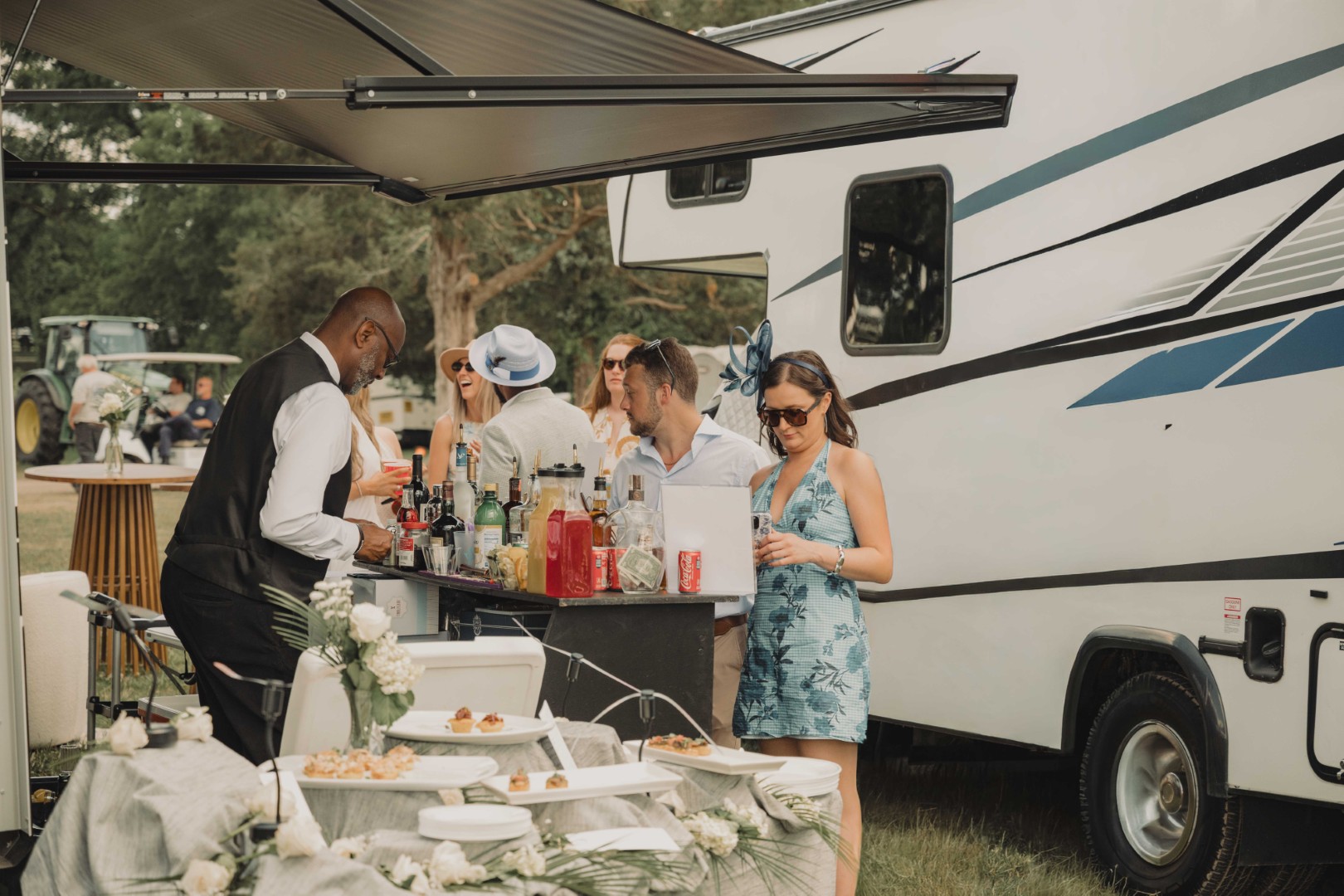 People gather by a bar setup next to a camper van with drinks and hors d'oeuvres outdoors.