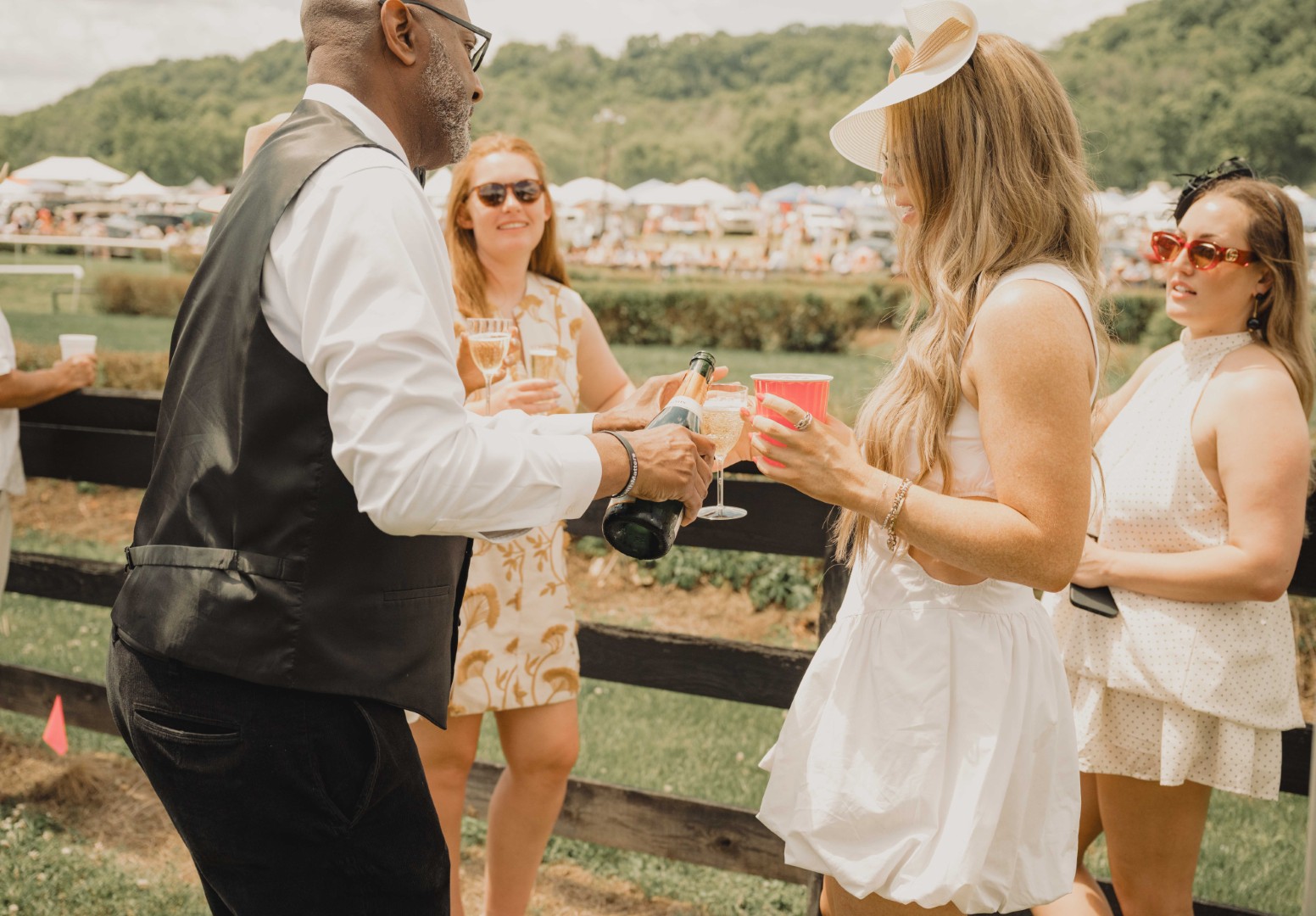 Man in vest pouring champagne for three women outdoors at a social event with a grassy field.