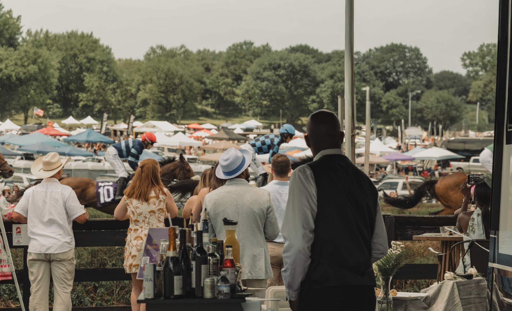 People watching a horse race with jockeys riding horses and tents in the background.