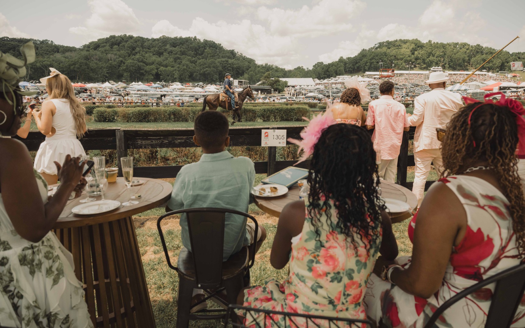 People at tables and standing watch a horseback rider at an outdoor event with tents and hills in background.