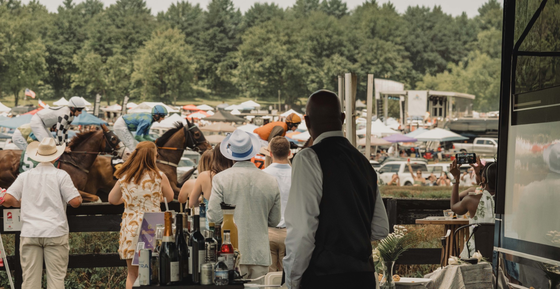 Crowd watching horse race with jockeys in colorful silks riding horses at an outdoor event.