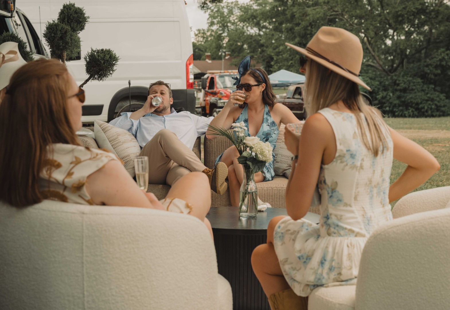 Four people relaxing and drinking on outdoor couches near a white van in a park setting.