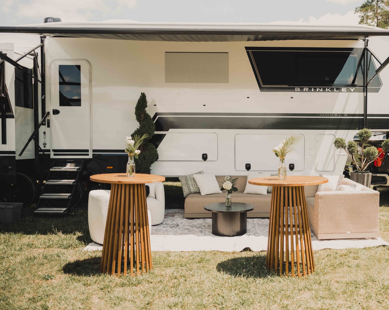 Outdoor seating area with a camper, two wooden tables, a couch, and potted plants on grass.