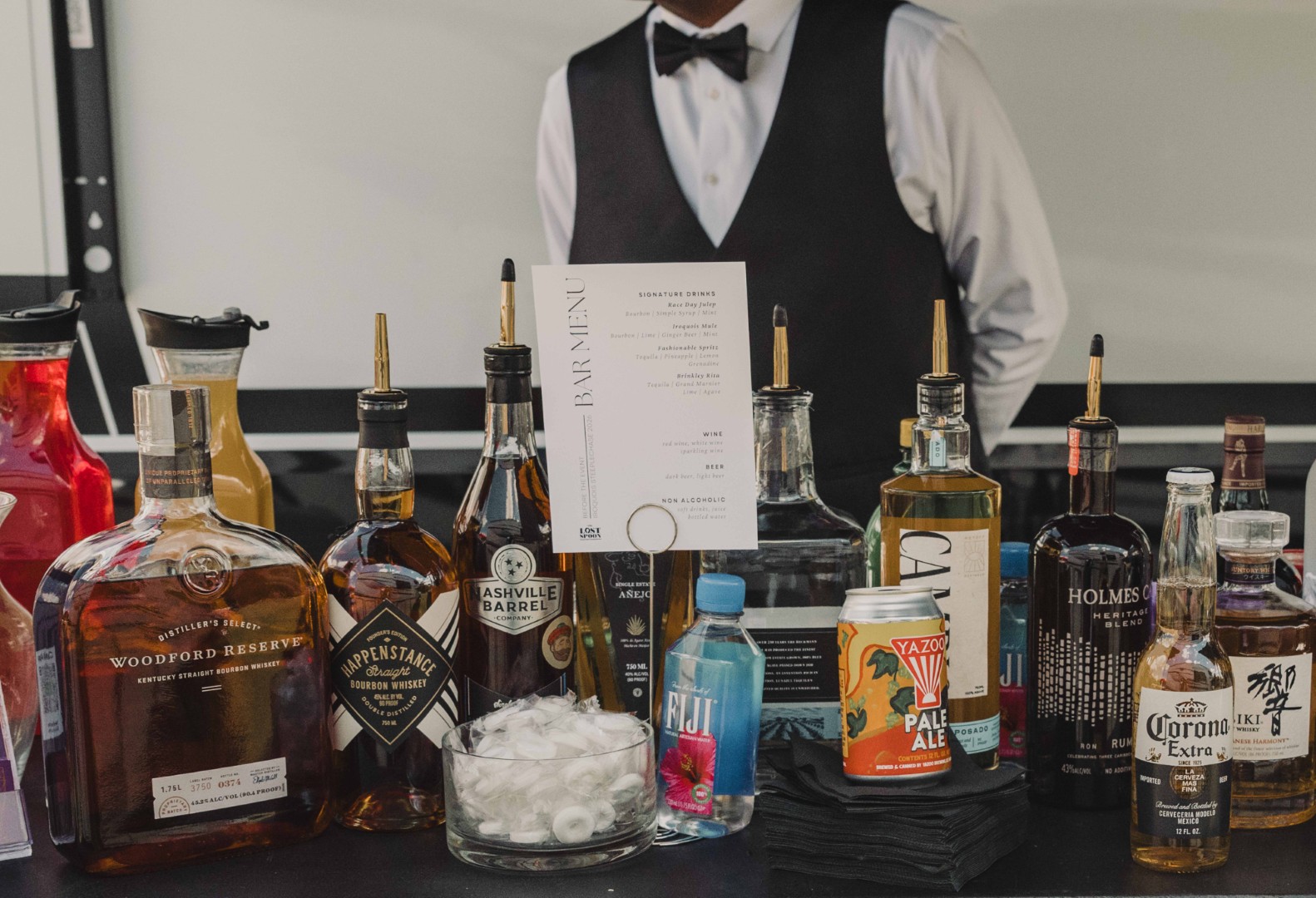 Bartender behind bar with assorted liquor bottles, a drink menu, ice bowl, and a bottled water on table.