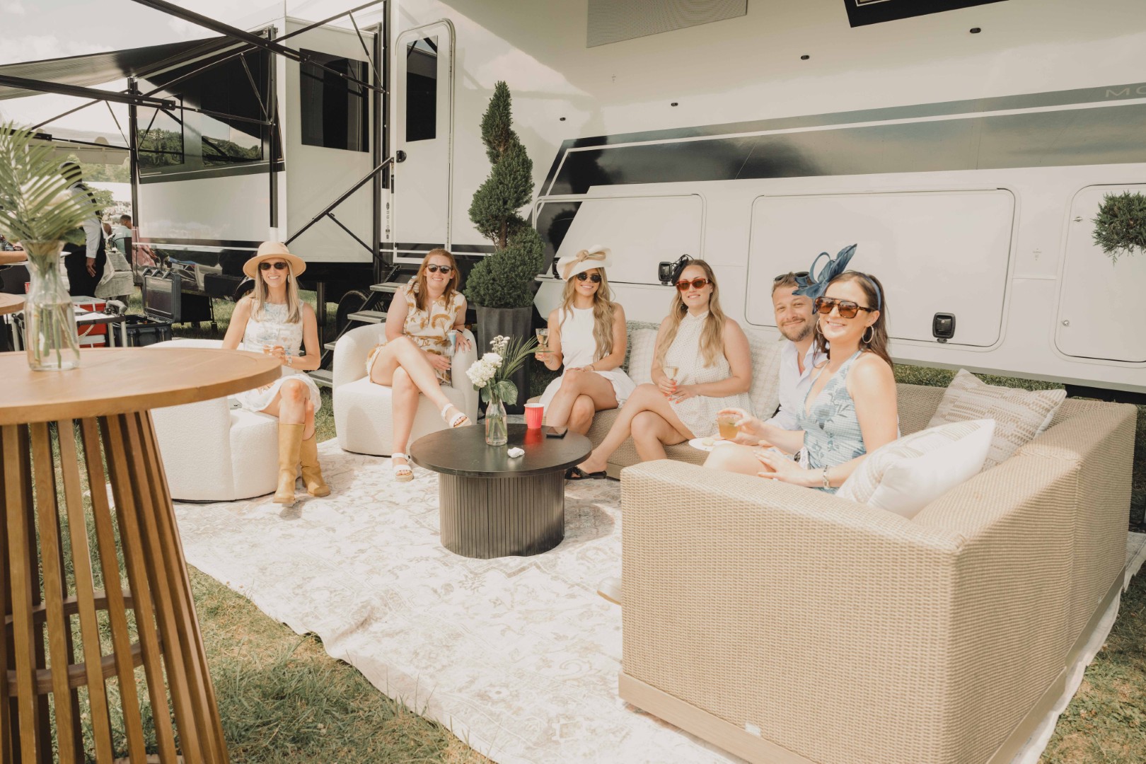 Six people in hats and sunglasses relaxing on outdoor sofas by a large white RV at an event.