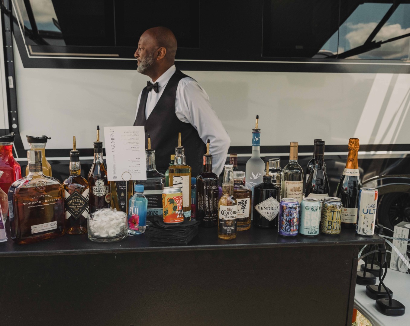 Bartender in vest stands behind a bar with various bottles of alcohol and canned drinks.