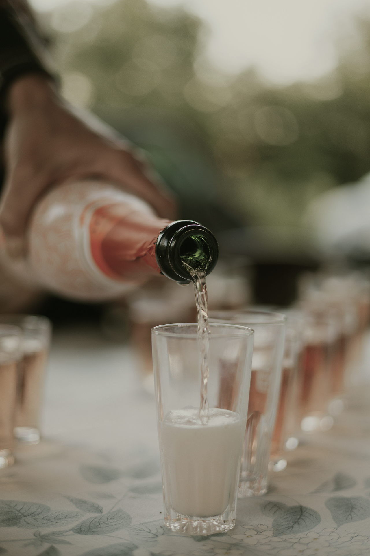 Hand pouring a light-colored drink from a bottle into a clear glass on a table with more glasses.