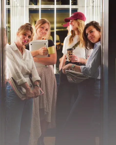 Quatre femmes souriantes tenant des tasses et des carnets dans un ascenseur moderne.