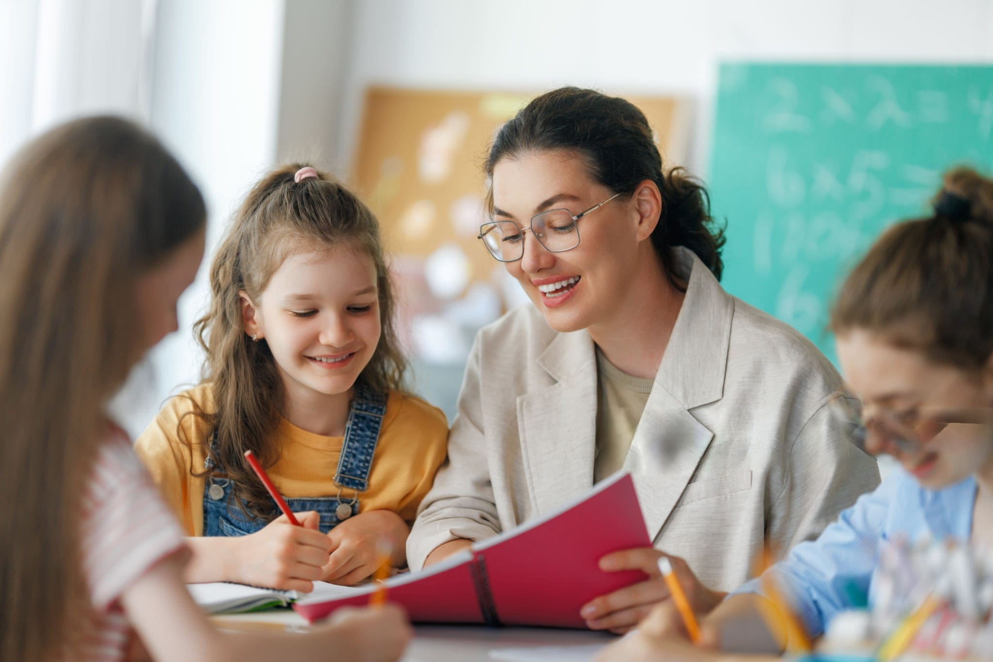 Respire Education | Teacher with glasses smiling and holding a notebook while helping a girl in a yellow shirt and denim overalls.