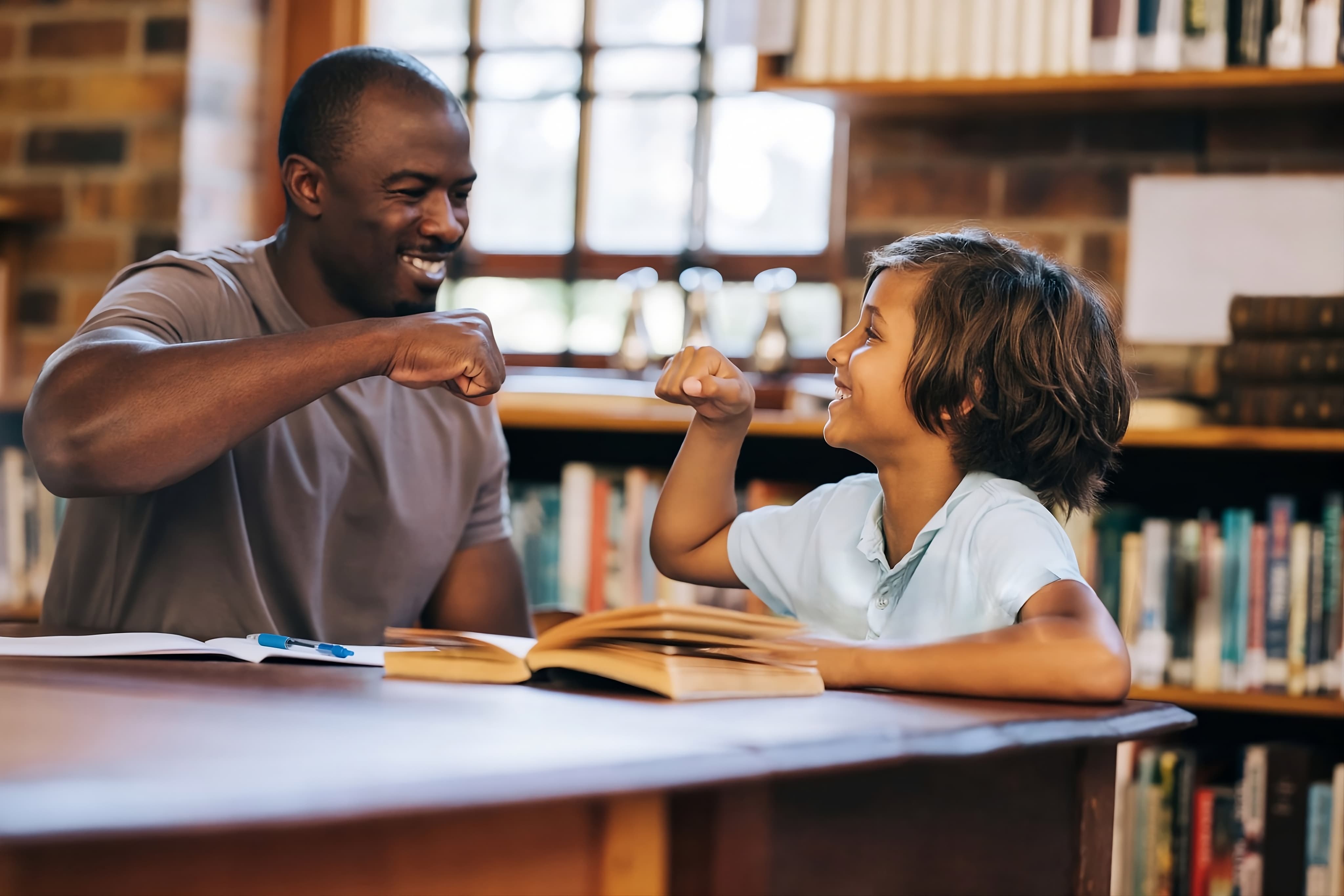 Respire Education | Smiling man and boy fist bumping across a library table with open books.