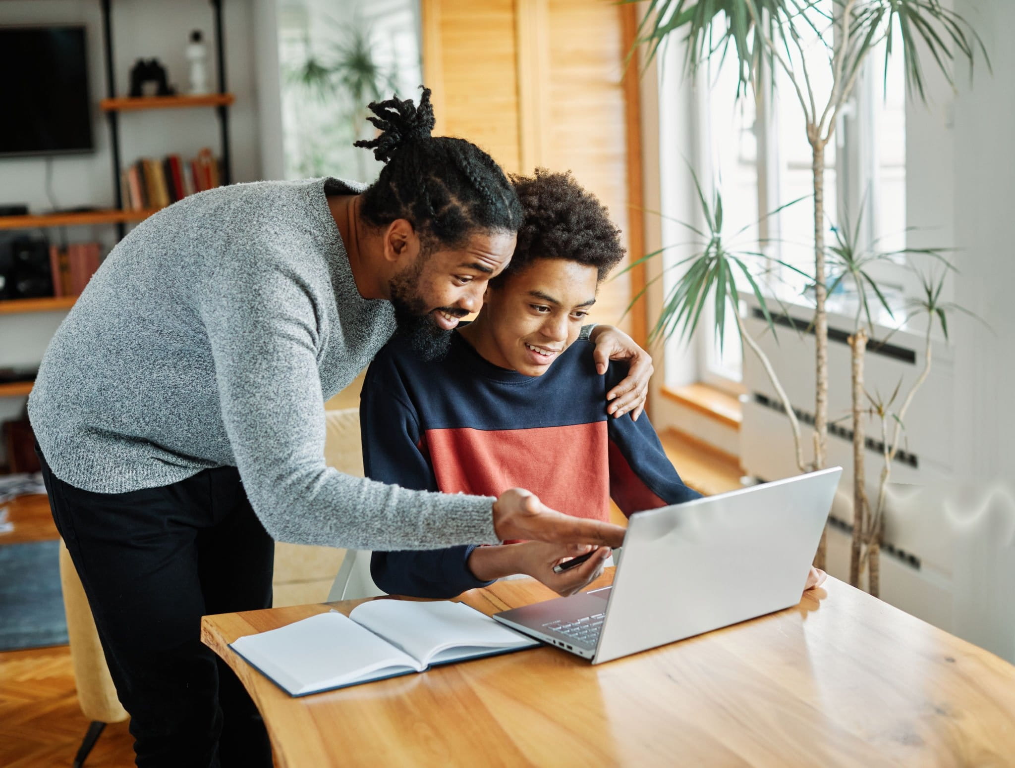 Respire Education | Man with dreadlocks helping smiling teenage boy with homework on laptop at wooden table.