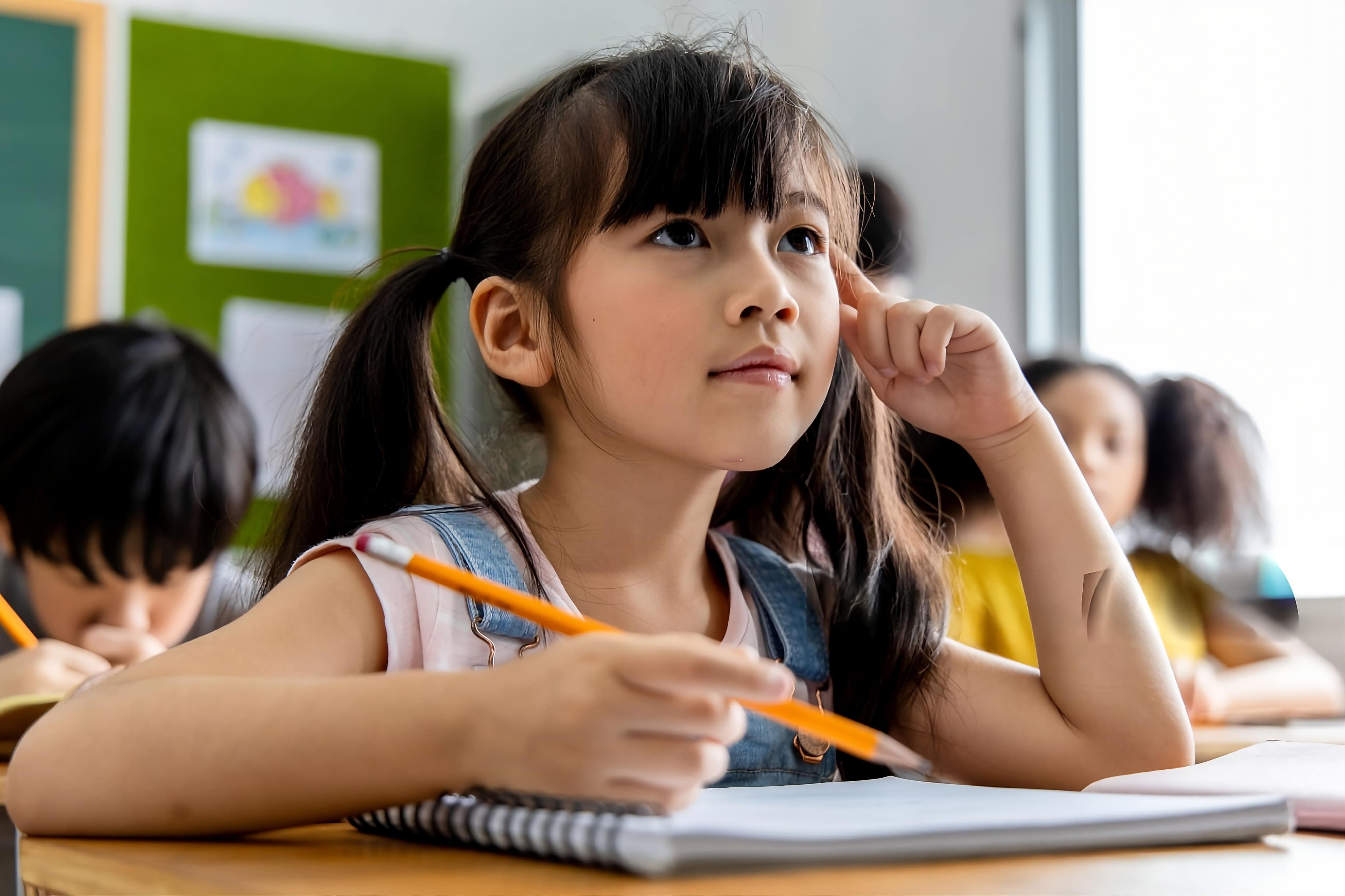 Respire Education | Young girl with pigtails holding a pencil and thinking in a classroom with other children.