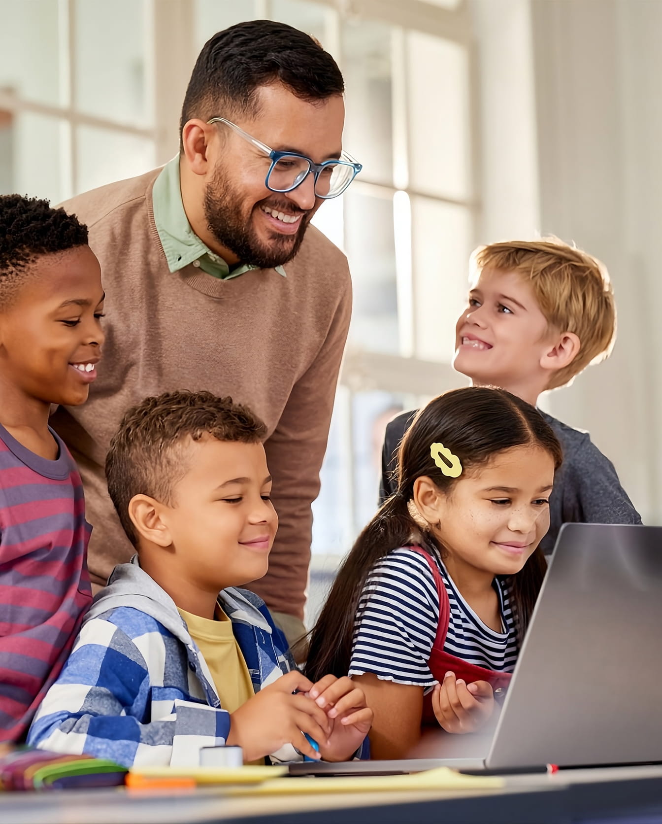 Respire Education | Teacher smiling with four diverse children gathered around a laptop in a bright classroom.