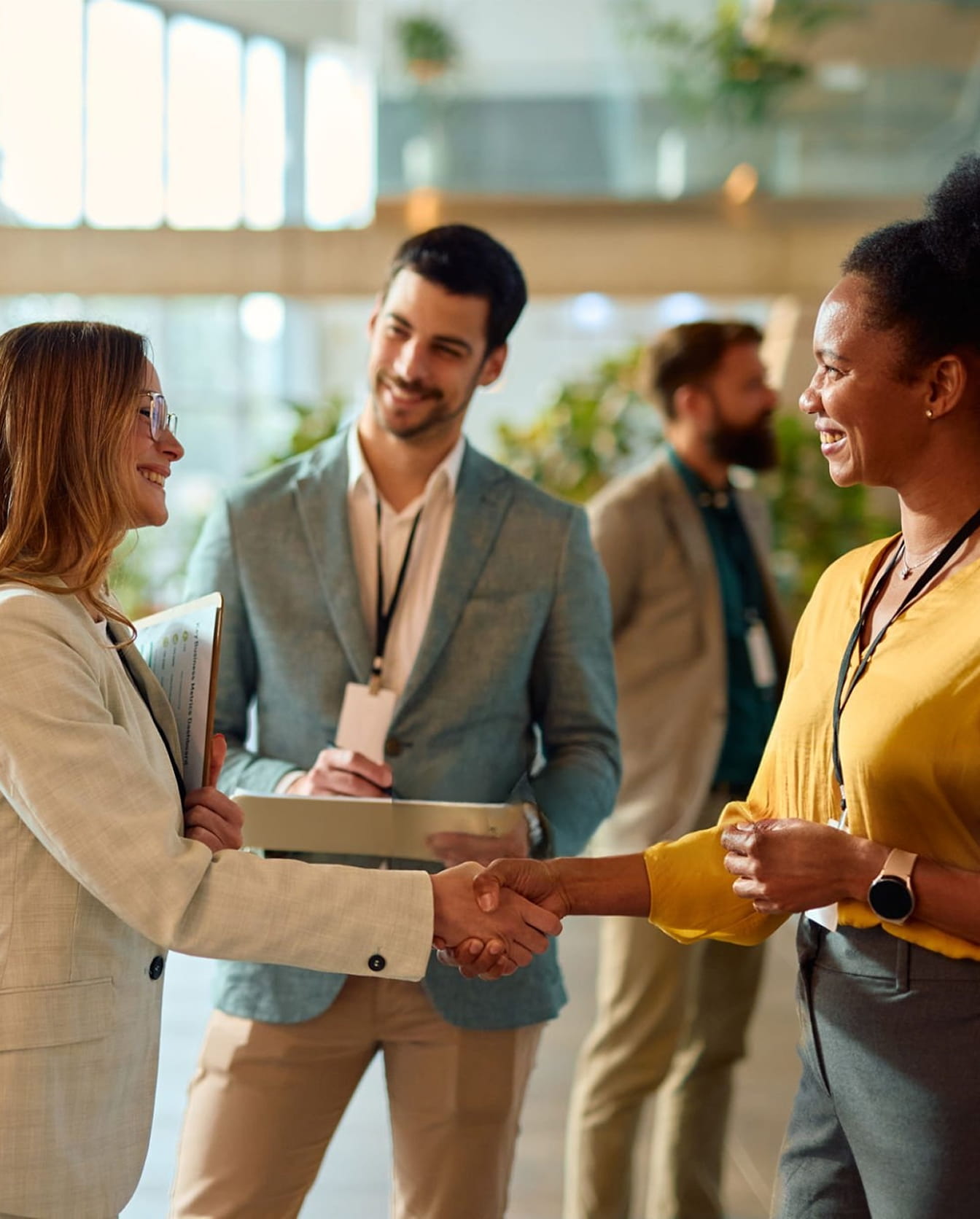 Respire Education | Two professional women smiling and shaking hands while two men observe in the background at a business event.