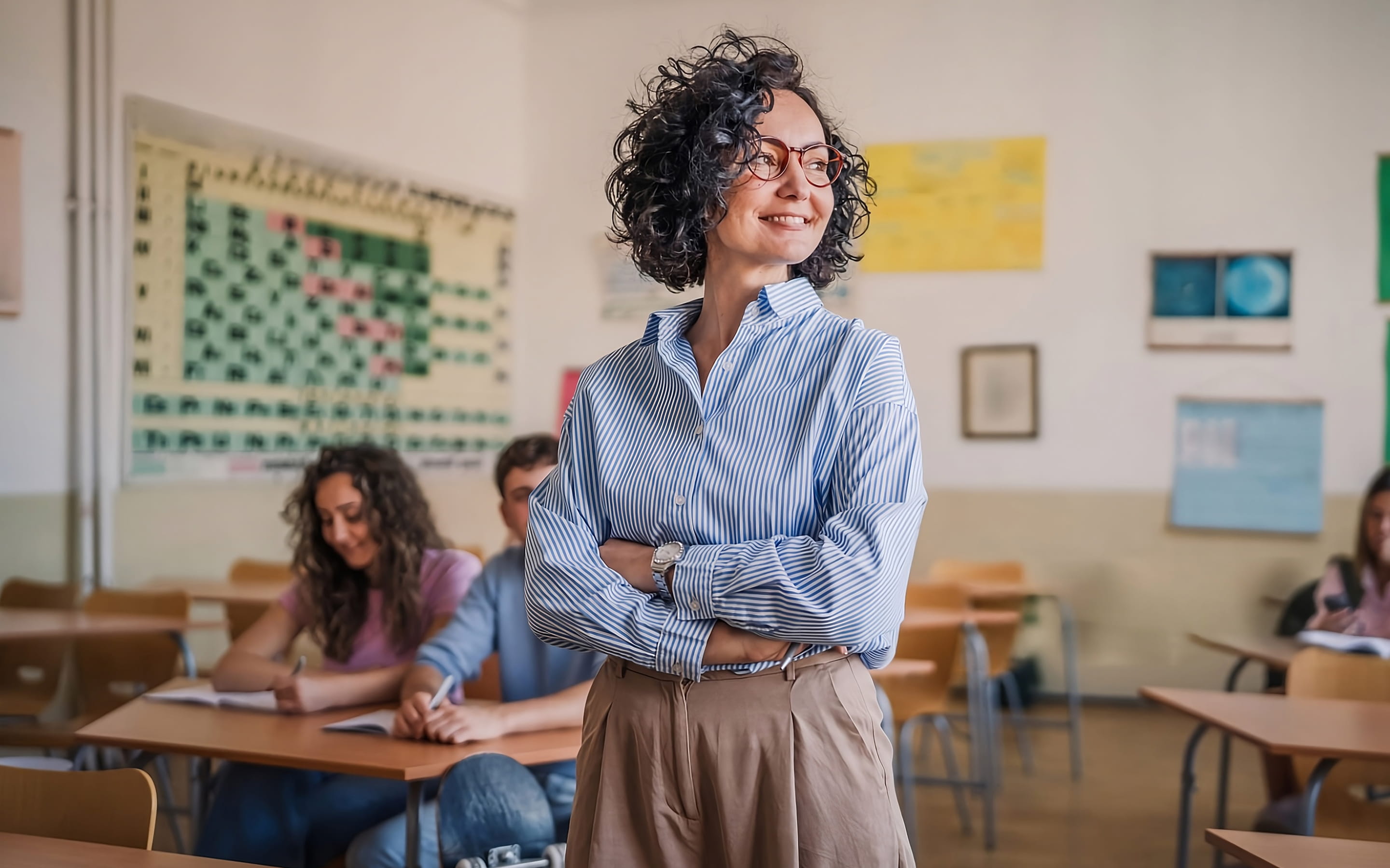 Respire Education | Confident female teacher with glasses standing in front of students in a classroom with a periodic table on the wall.