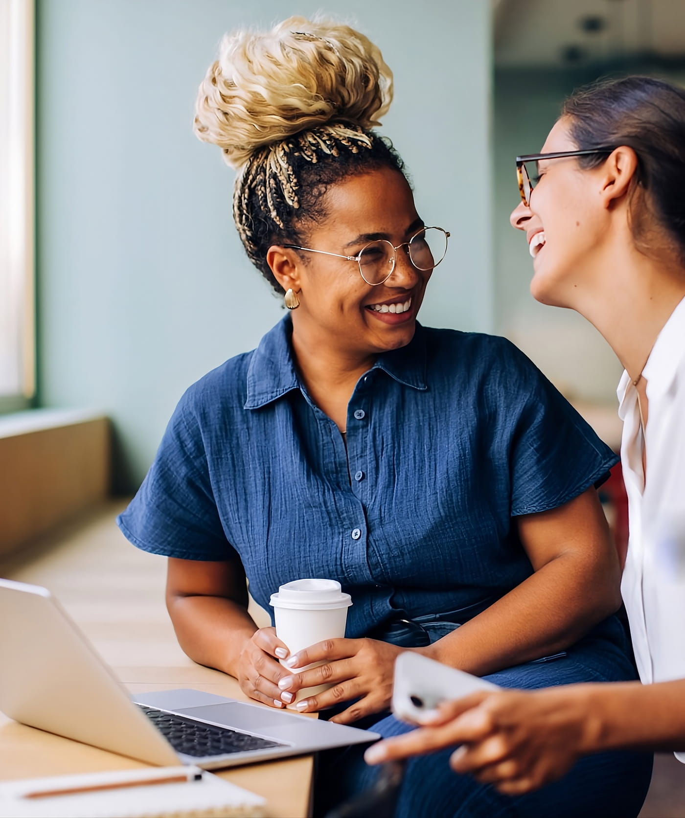 Respire Education | Two women smiling and chatting near a laptop, one holding a coffee cup and the other holding a phone.