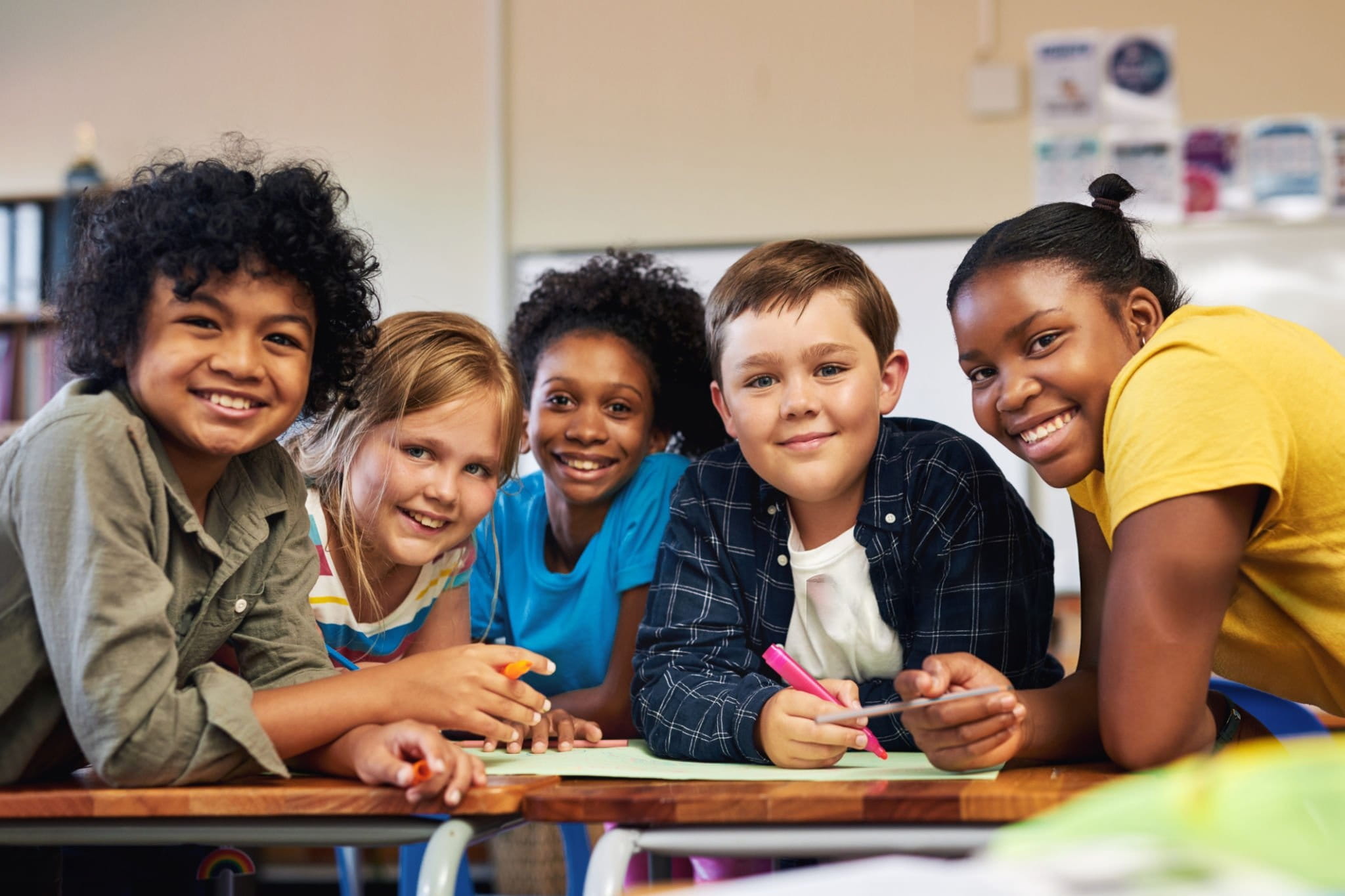Respire Education | Five diverse children smiling together at a table in a classroom while holding markers.