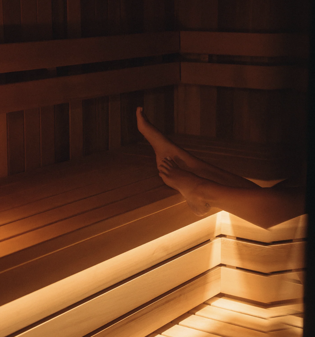 Person's bare legs resting on a wooden bench inside a softly lit sauna room.