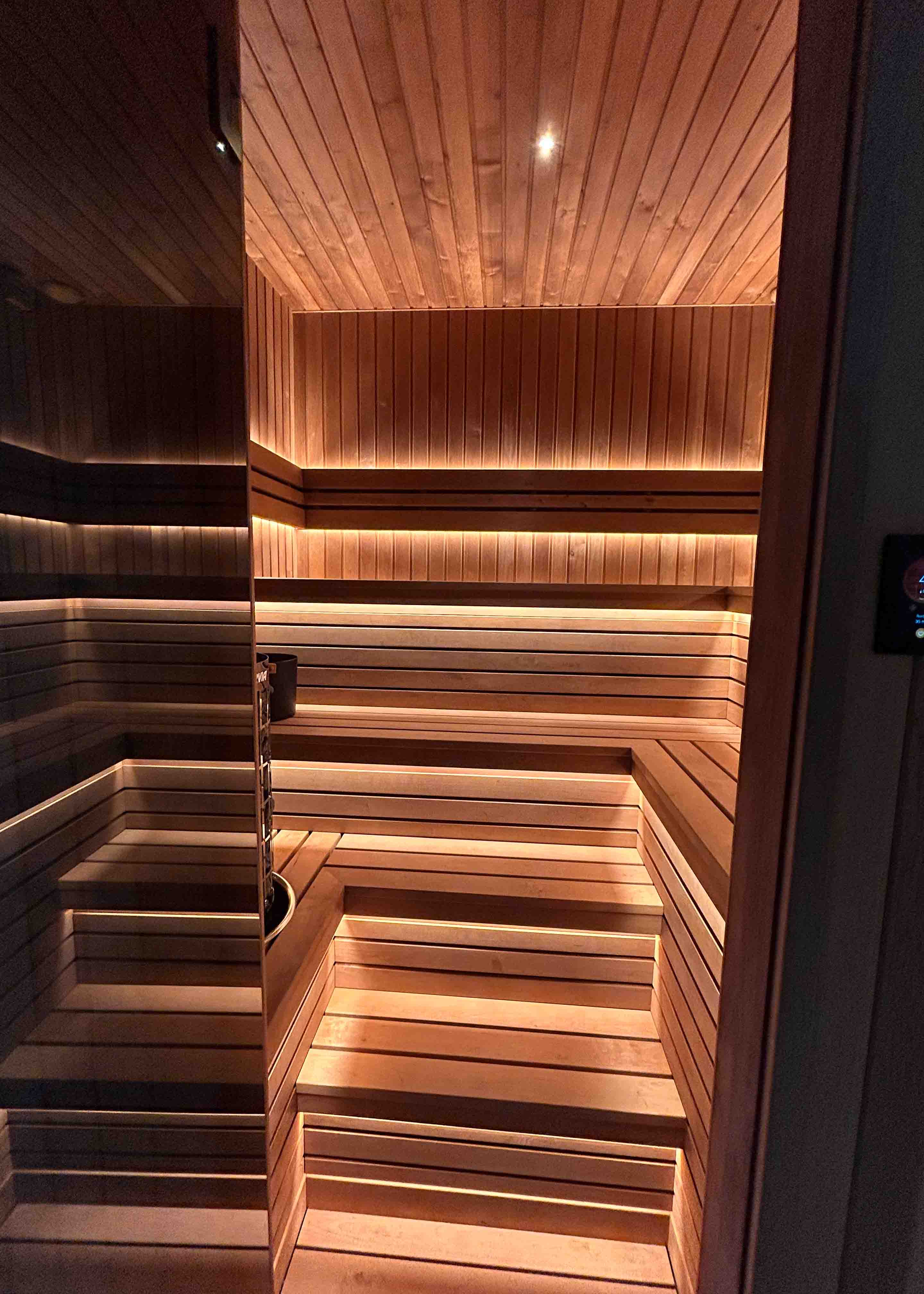 Interior of a wooden sauna with tiered benches, backrest, lighting, and a heater with sauna stones.