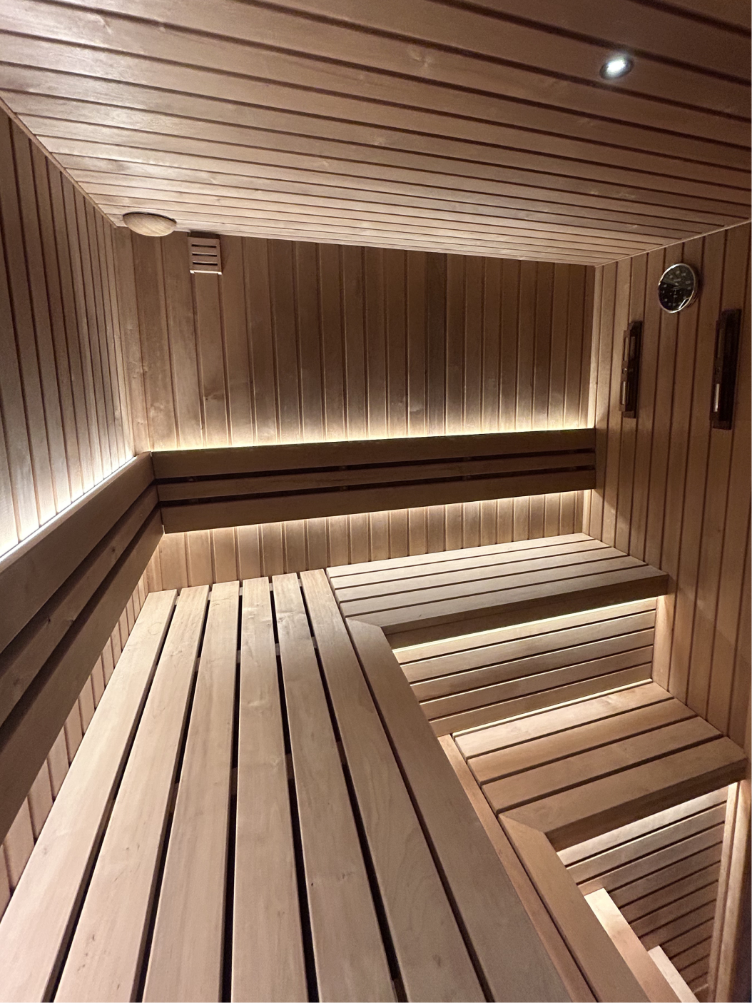 Interior of a wooden sauna with tiered benches, backrest, lighting, and a heater with sauna stones.