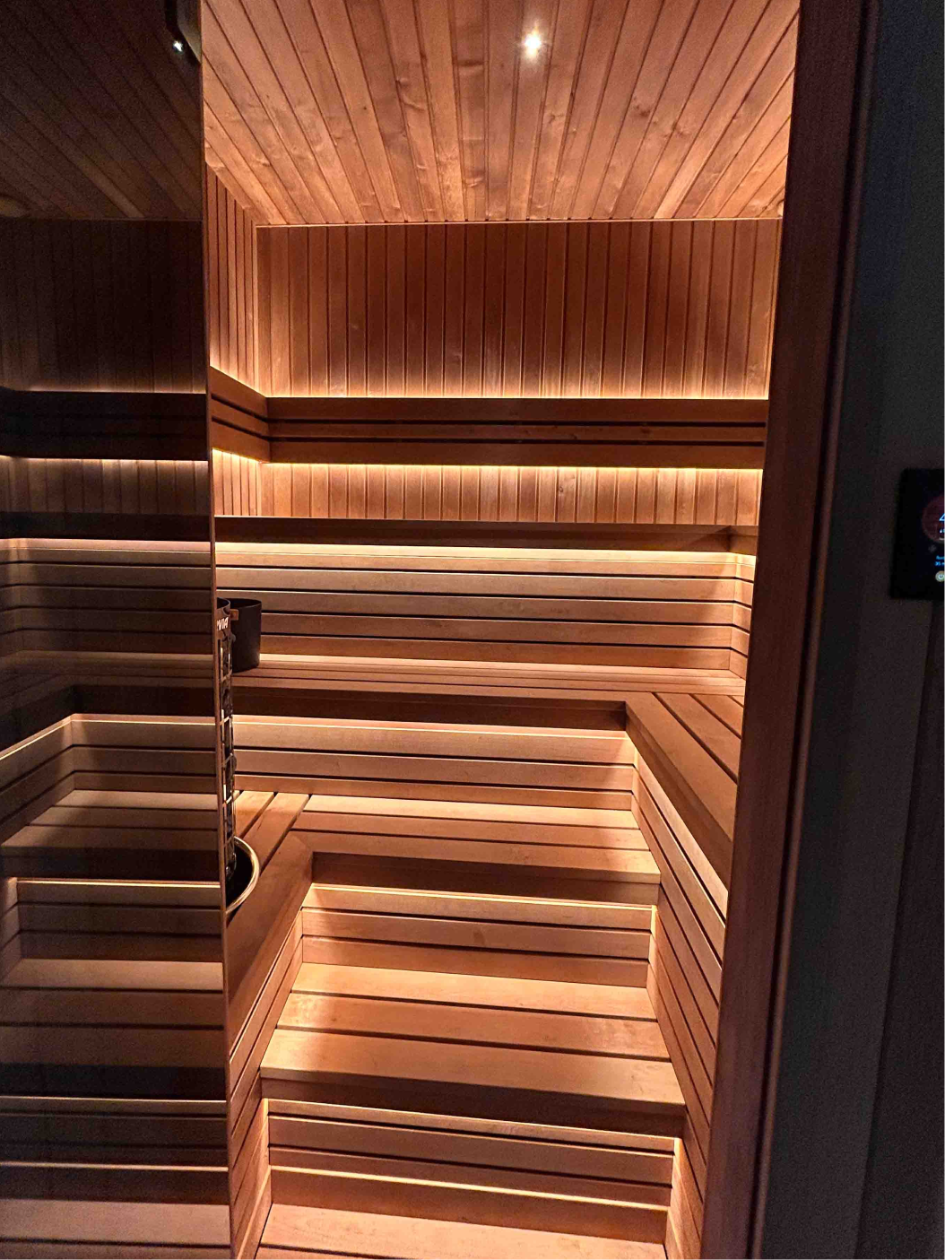 Interior of a wooden sauna with tiered benches, backrest, lighting, and a heater with sauna stones.