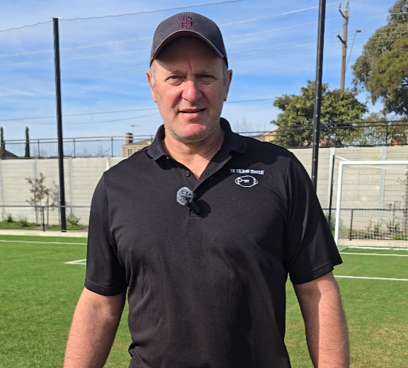 Man wearing a black polo shirt and cap standing on a green sports field with a goalpost behind him.