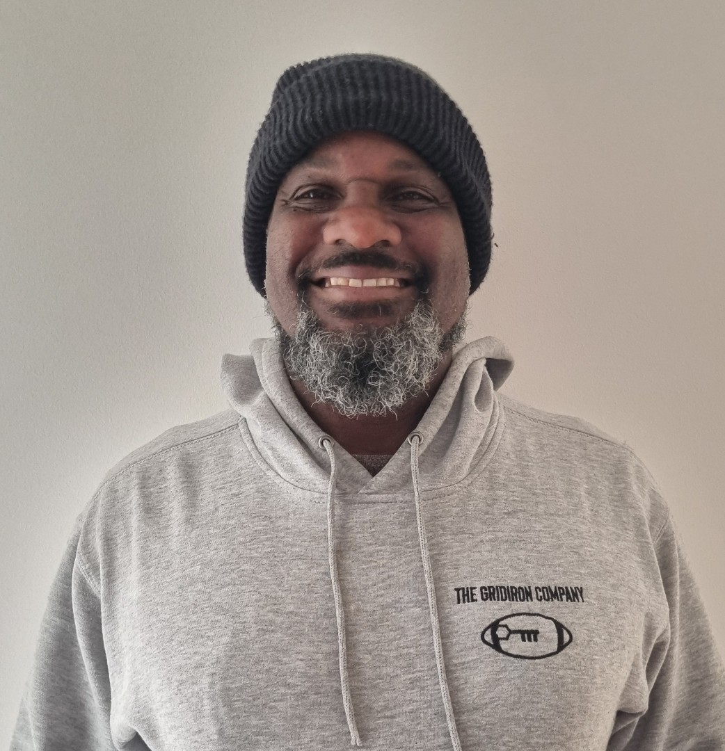 Smiling man with grey beard wearing black beanie and grey hoodie with The Gridiron Company logo.