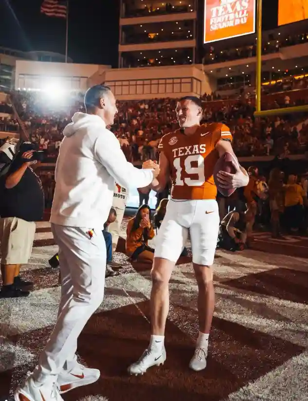 Texas football player in orange jersey holding ball shaking hands with man in white hoodie on field.