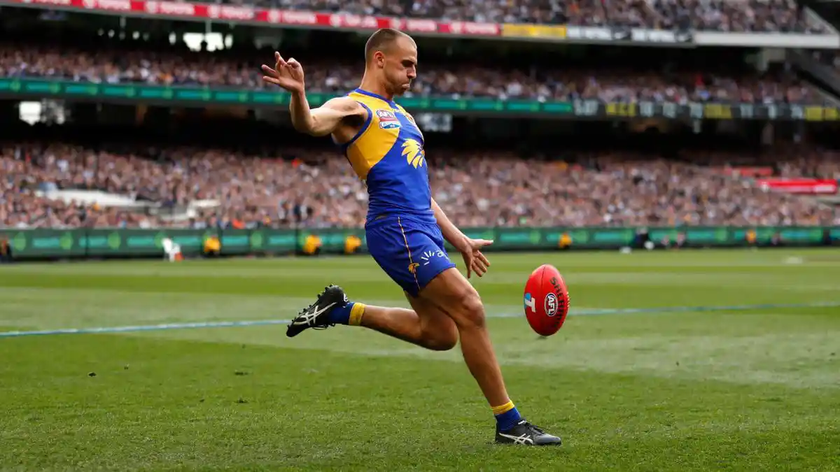 Australian football player in blue and gold kicking a ball in a stadium with a large crowd.