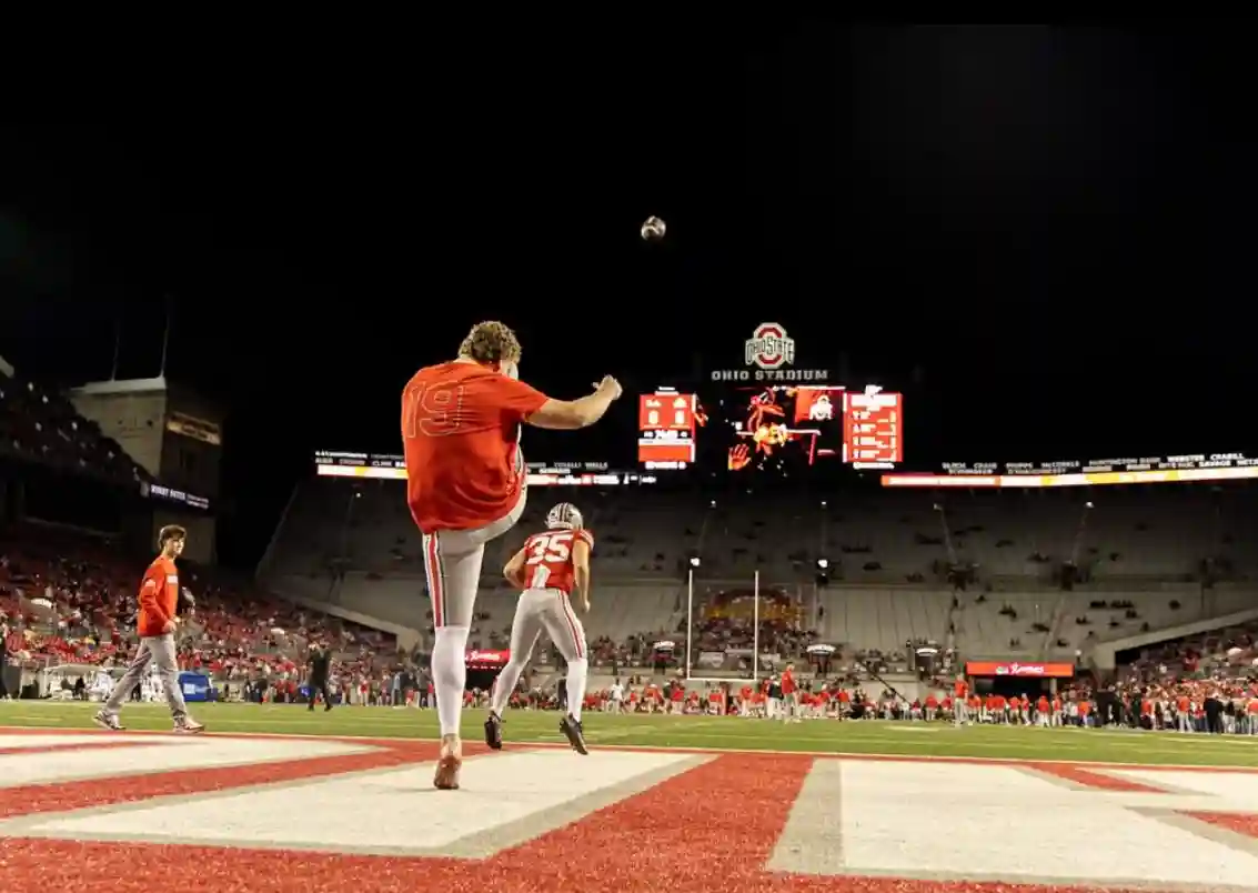 Football player in red jersey kicking ball in end zone at Ohio Stadium during game night.