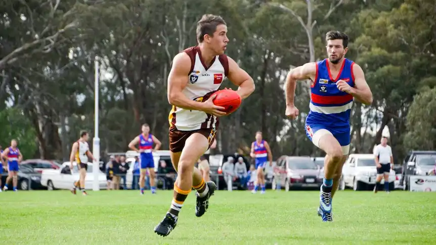 Australian rules football player in white and brown runs with ball while opponent in blue chases on grass field
