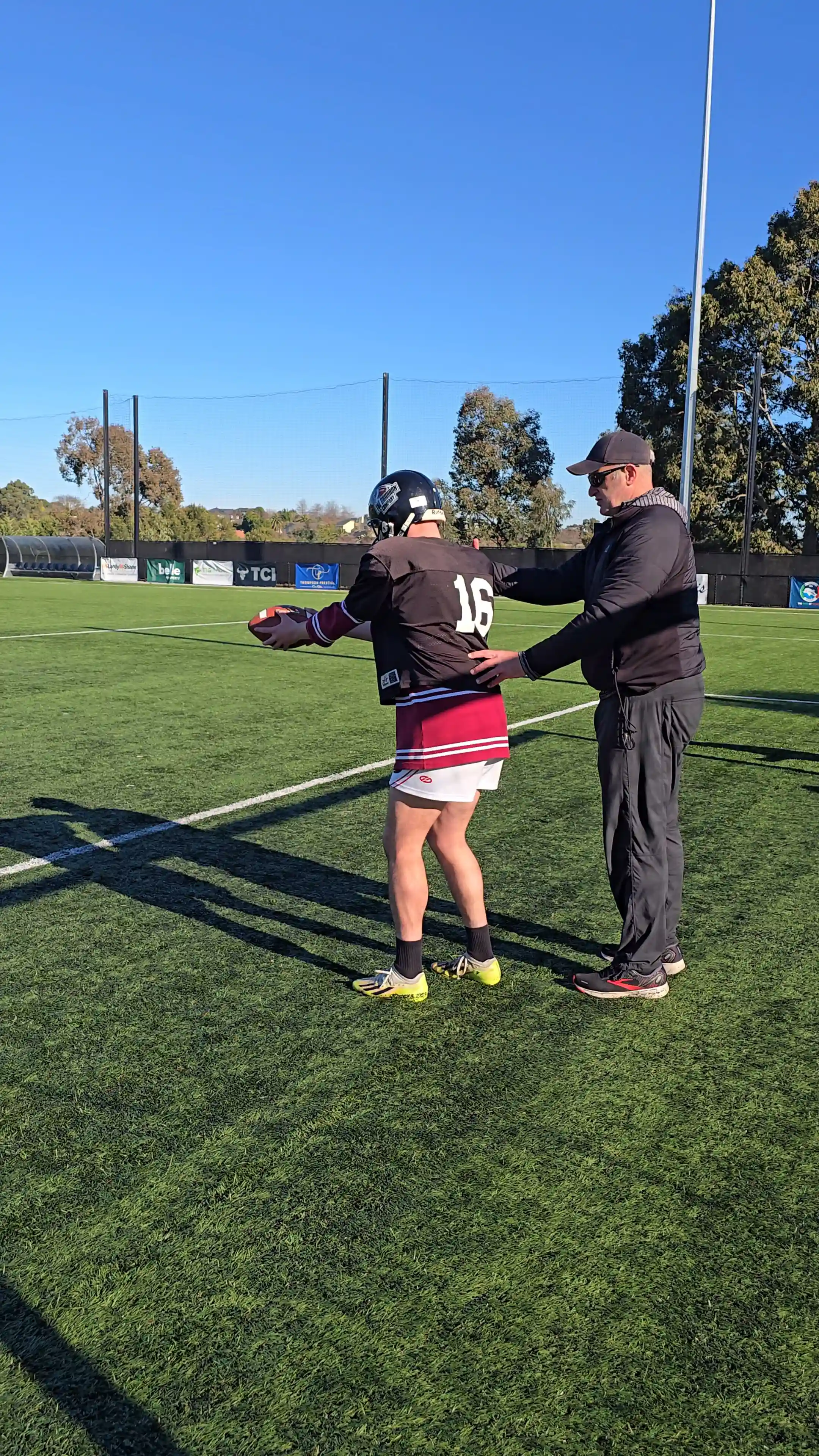 Coach guiding a football player holding a ball on a green field under a clear blue sky.