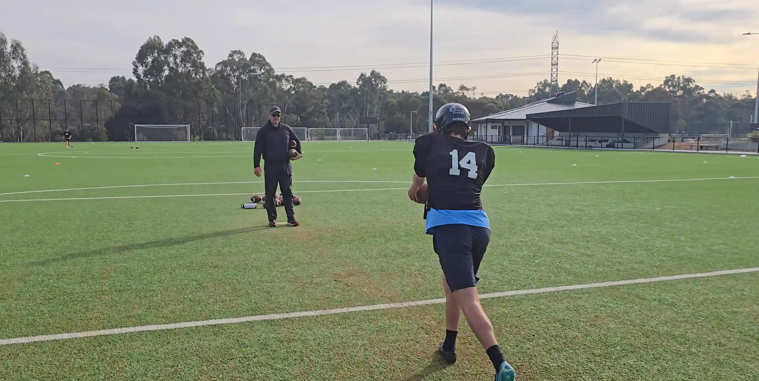 Young player in black #14 runs on a green field passing a football to a man in black with cap.