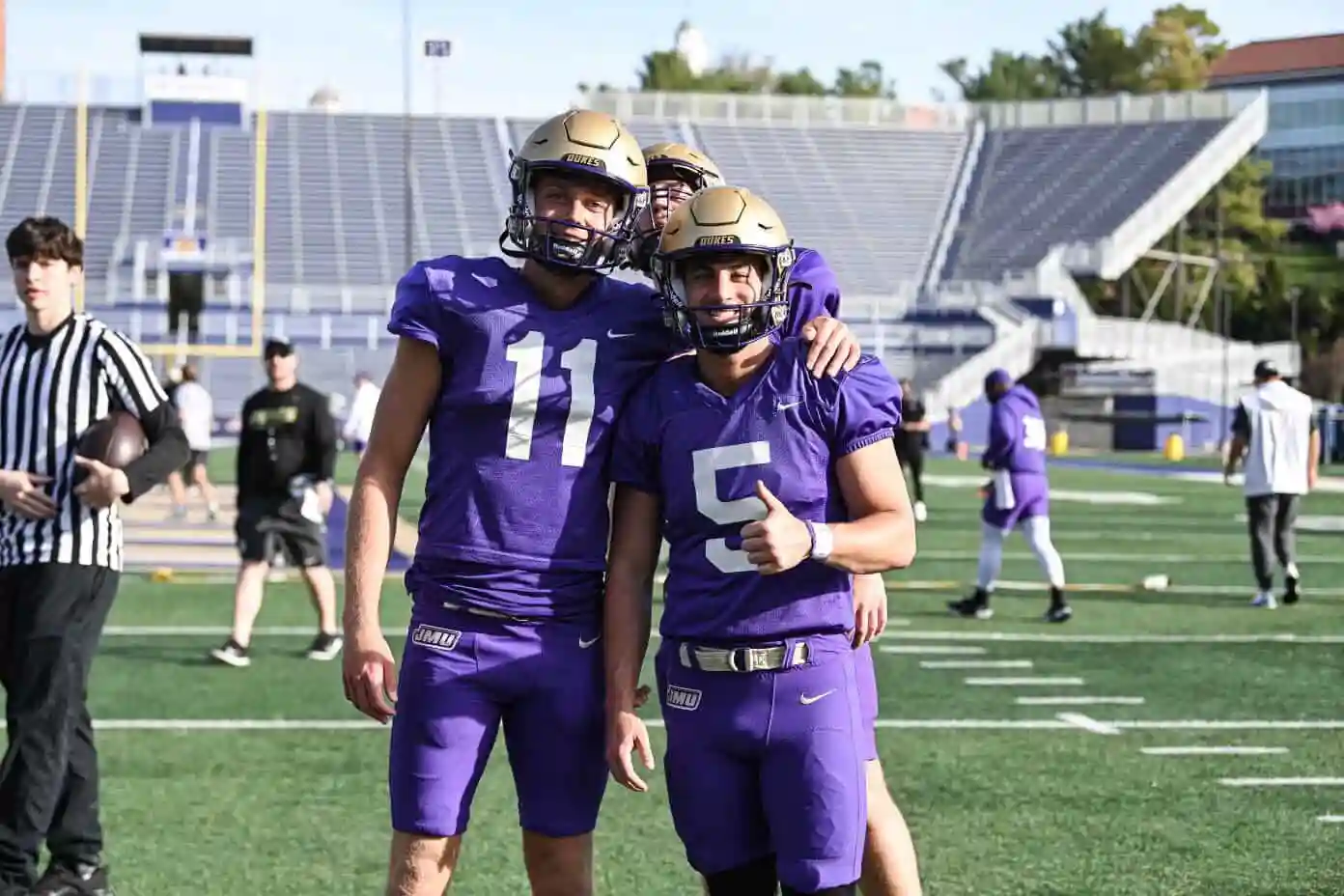 Three football players in purple uniforms and gold helmets posing on a green field in a stadium.