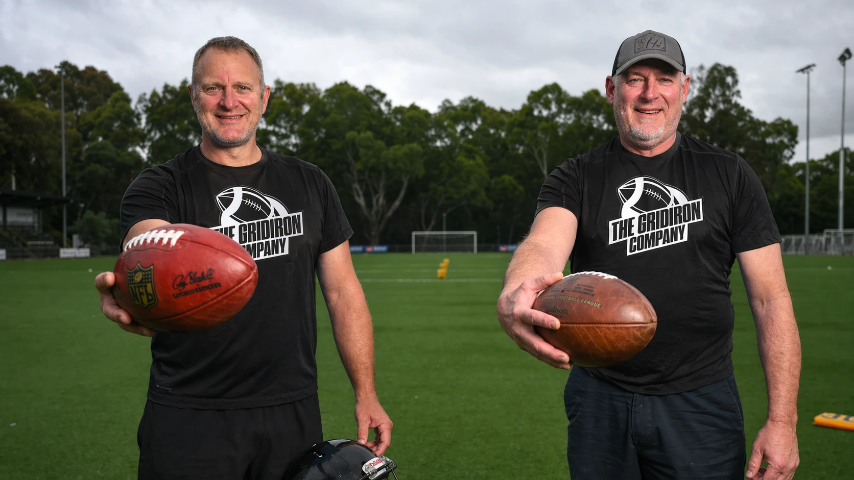 Two men wearing The Gridiron Company t-shirts holding footballs on a green sports field.